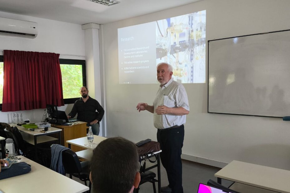El Lic. Javier Díaz y el prof. Agustín Candia en el aula de posgrado de la Facultad de Informática. Sobre la pared se proyecta una imagen y un pizarrón.