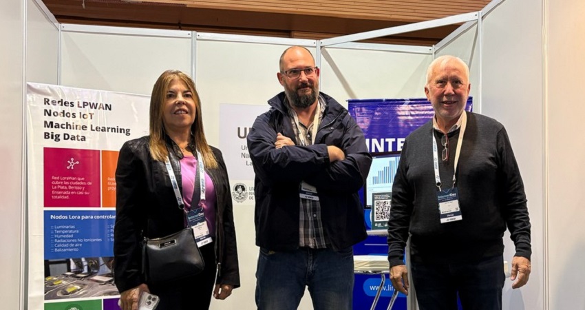 Tres personas de pie en un stand de exposición: a la izquierda una mujer con saco negro y blusa fucsia, en el centro un hombre con barba, anteojos y chaqueta oscura con los brazos cruzados, y a la derecha un hombre mayor de cabello cano y suéter negro. Detrás se observan carteles del LINTI/CeSPI con textos sobre LPWAN, nodos IoT, machine learning y una pantalla con gráficos; el ambiente corresponde al Internet Day 2026